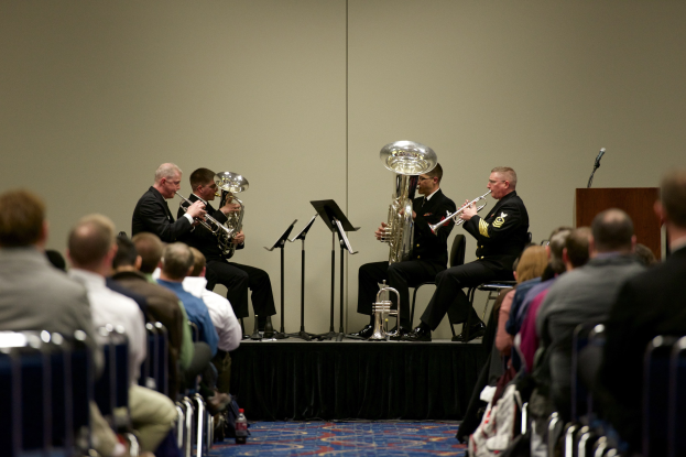 Eine Gruppe von Männern, die auf einer Bühne Blasinstrumente spielen, mit Notenständern, einem Podium mit Mikrofon und einer Sitzreihe im Publikum, mit einer Wand im Hintergrund.