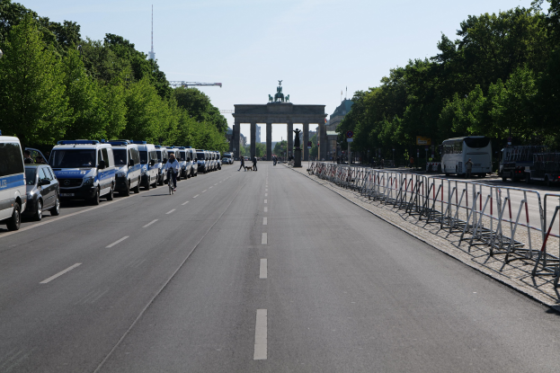 Lange Reihe von Polizeiwagen, die entlang einer Straße vor dem Brandenburger Tor geparkt sind, mit Fahrradfahrern und Fußgängern in der Nähe, Absperrungen, Bäumen und einem prächtigen Bogen im Hintergrund.
