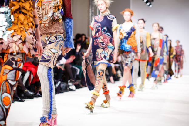 Eine Gruppe Von Frauen, Die Eine Laufsteg In London Fashion Week Heruntergehen, Mit Einem Publikum Im Hintergrund Unter Helleren Lichtern.