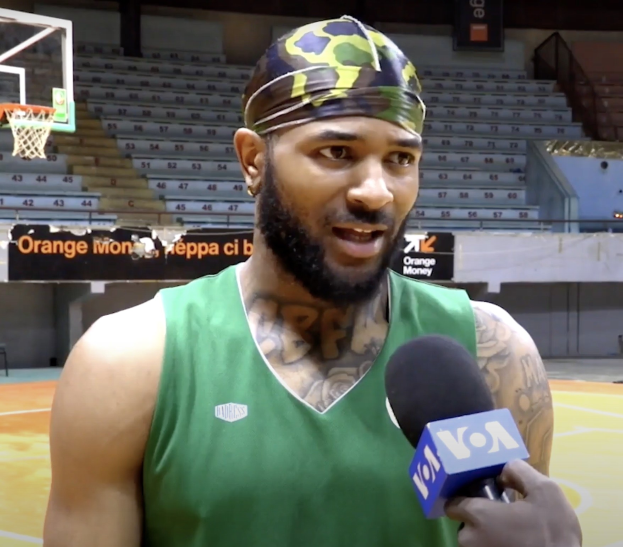 Mann in grüner Tanktop, der vor einem Basketballfeld mit Netz auf der linken Seite und Treppen im Hintergrund in ein Mikrofon spricht.