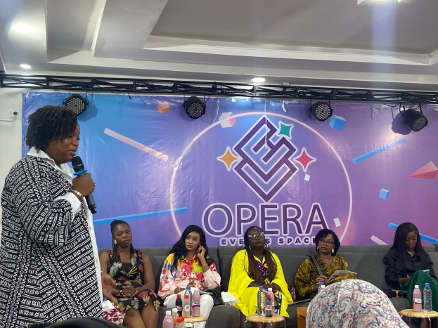 Frau steht vor sitzenden Frauen auf einer Couch, hält ein Mikrofon, mit Flaschen und Gegenständen auf den Tischen im Vordergrund und einem Banner mit Text und Deckenbeleuchtung im Hintergrund.