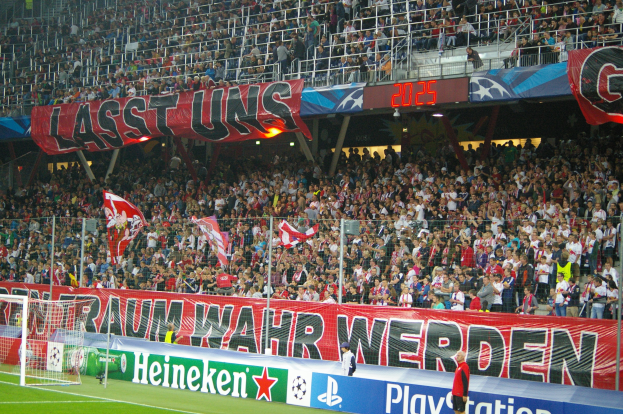 Fußballspiel in einem Stadion mit großer Zuschauermenge, saftigem Grün, einem Torpfosten links, Bodenbanner, Menschen mit Fahnen und Hintergrundgeländer, Tafeln, Lampen und andere Gegenstände.