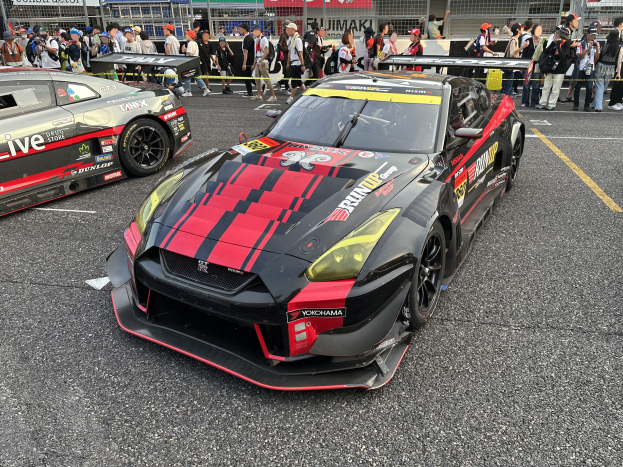 Ein Nissan GT-R Nismo GT3-Rennwagen auf der Straße, umgeben von einer Menschenmenge, einige machen Fotos, mit Gebäuden, Texttafeln, Polen und einem klaren blauen Himmel im Hintergrund.
