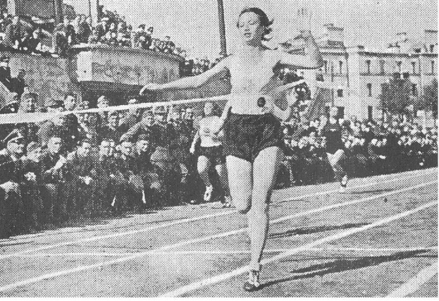 Schwarzes und weißes Foto einer Frau beim 100m-Finale bei den Olympischen Spielen 1956 auf einer Laufbahn mit Zuschauern, Gebäuden und Pfosten im Hintergrund.