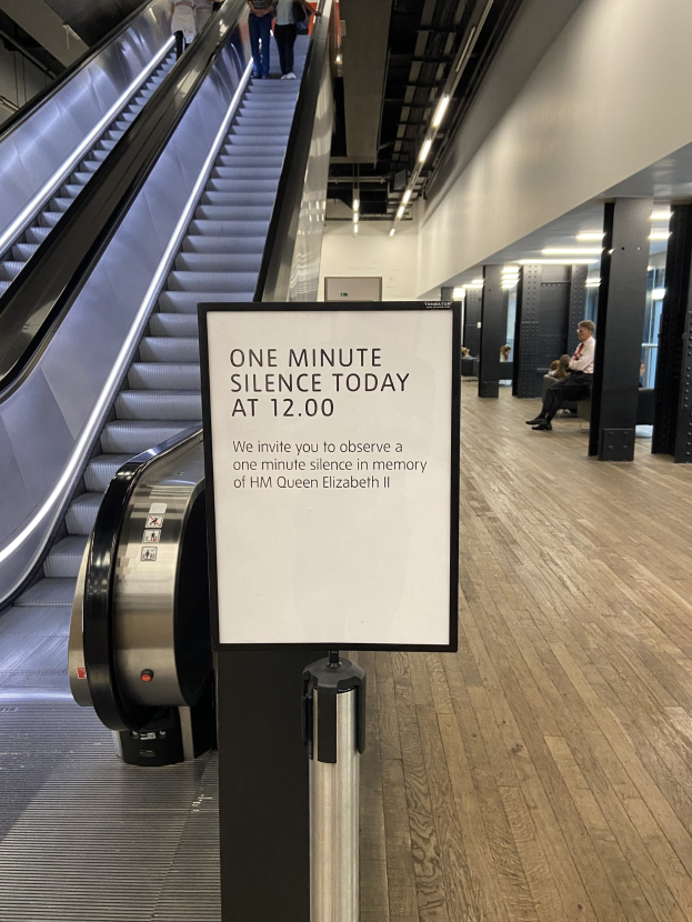 Eine Rolltreppe in einem Flughafen mit einer Tafel, auf der "Eine Minute Stille heute" steht, sowie ein paar Menschen darauf und an der Decke angebrachte Lichter im Hintergrund.