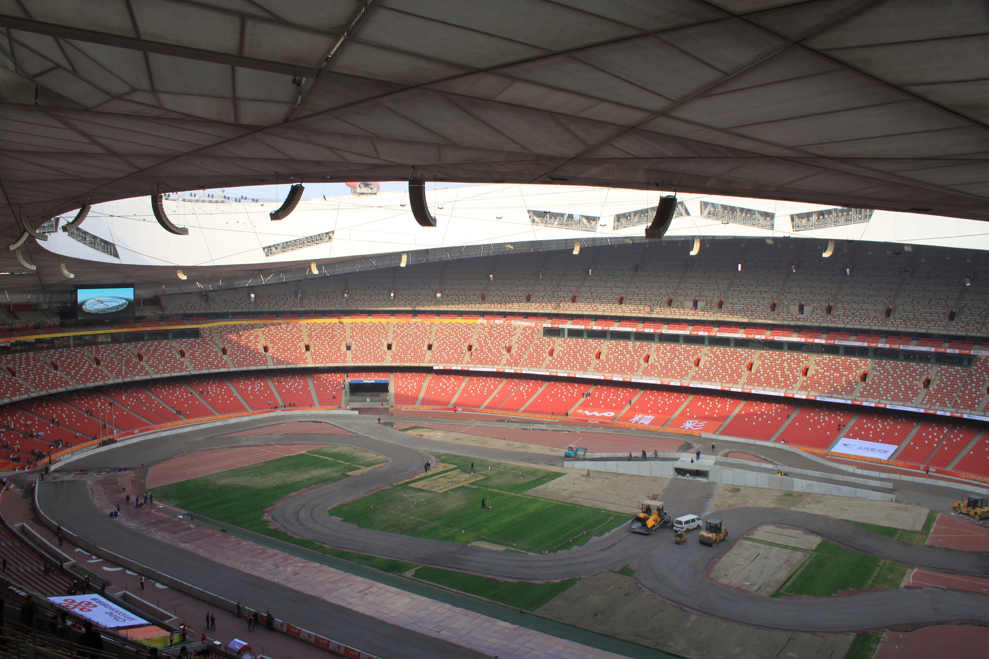 Ein großes Stadion voller Menschen, mit Fahrzeugen auf der Straße, Gras, Texttafeln, Geländern, einem Bildschirm und Deckenleuchten.