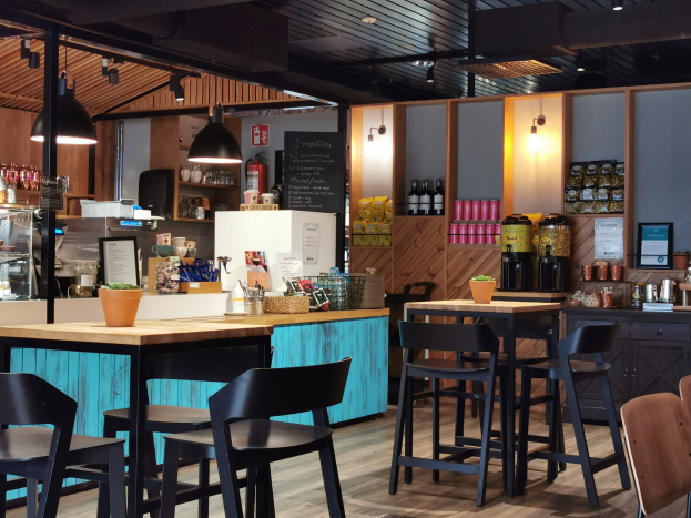 Innenraum eines Restaurants mit Holztischen, Stühlen, einer Topfpflanze auf einem Tisch, einer Schiefertafel, einem Kühlschrank, einem Feuerlöscher, Regalen mit Flaschen, Schränken mit Gegenständen, einem Rahmen mit einem Foto an der Wand und Deckenleuchten.