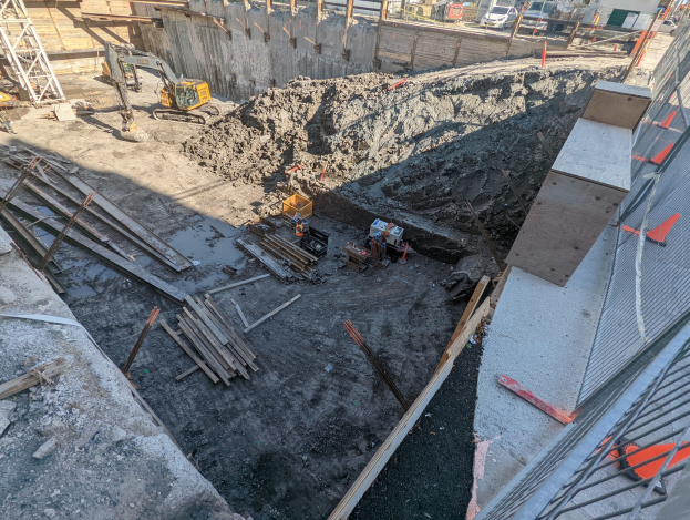 Baustelle mit einem großen Haufen Erde, umgeben von Kränen, Holzstöcken und anderem Equipment, begrenzt von einem Maschendrahtzaun auf der rechten Seite, mit Gebäuden, Fahrzeugen, Pfählen, Bäumen und Himmel im Hintergrund.