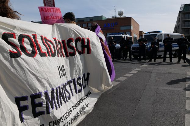Eine Gruppe von Menschen marschiert mit einer 'Solidarität und Feminismus'-Schlagzeile die Straße entlang, mit geparkten Fahrzeugen, Gebäuden, einer Schüsselantenne und einem klaren blauen Himmel im Hintergrund.