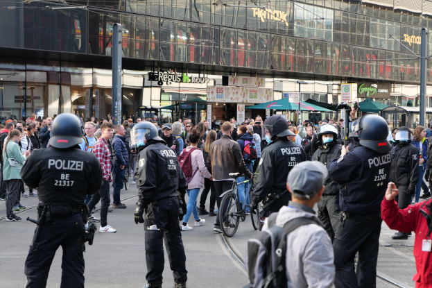 Eine Gruppe von Polizisten steht vor einer Menschenmenge auf der Straße, einige tragen Helme und halten Fahrräder, im Hintergrund Gebäude mit Glaswänden und Schildern.