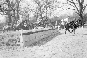 Schwarzes und weißes Foto von Menschen, die Pferde über eine grüne Wiese mit einer Wand und Stangen im Vordergrund reiten, Bäume und Himmel im Hintergrund und Text unten, der "Pferdespringen bei der National Horse Show in Washington, D.C. USA" lautet.