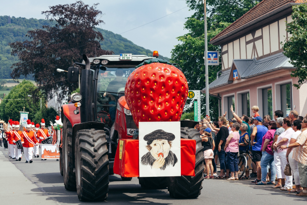 Ein Traktor mit einer großen Erdbeer-Dekoration fährt eine Straße entlang, während eine Gruppe von Menschen mit Fahrrädern auf dem Bürgersteig steht; Gebäude, Bäume und Hügel sind im Hintergrund unter einem klaren blauen Himmel zu sehen.
