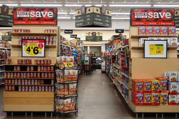 Ein Kiosk-Innenraum mit verschiedenen Lebensmitteln auf Regalen, Regalen und dem Boden, Deckenleuchten und Informationsschildern im Hintergrund.