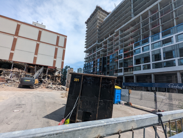 Großes Gebäude mit Schutt davor, umgeben von einem Metallzaun, einem Container, Pflanzen, Bäumen, Pfählen, einem Schild, Fahrzeugen, Steinen und einem bewölkten Himmel auf einer Baustelle für einen neuen Wohnkomplex.