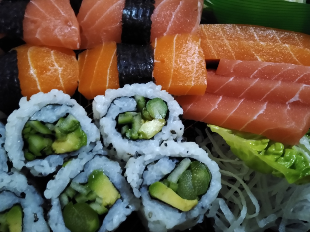 Ein schwarzer Teller mit verschiedenen Sushi-Rollen, darunter Lachs, Avocado und Gurke, umgeben von ein paar grünen Blättern.