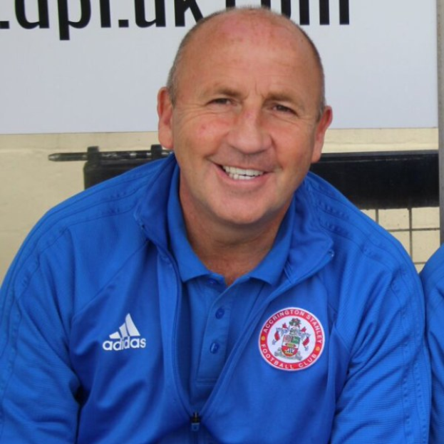 Zwei lachende Männer in einer blauen Jacke und ein weiterer Mann sitzen neben einer Wand mit einer Tafel, wobei der Mann in der blauen Jacke wahrscheinlich der Trainer von Accrington Stanley Football Club ist.