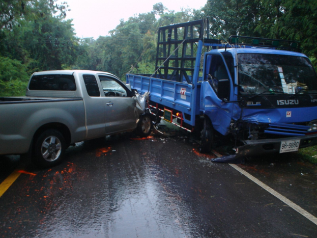 Ein schwerbeschädigtter Lkw mit eingedrückter Front und verbeulter Karosserie liegt auf der Seite einer Straße, umgeben von Bäumen unter einem klaren blauen Himmel.