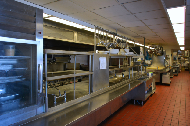 Gewerbeküche mit Edelstahl-Theke, aufgehängten Utensilien, einem Spülbecken, Deckenbeleuchtung, Schränken und verschiedenen Gegenständen auf Regalen.