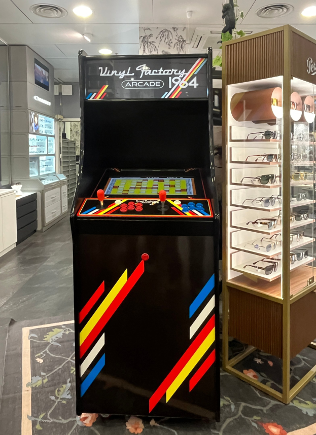 Ein Retro-Laden mit einem alten Arcade-Automaten in der Mitte, einem Gestell mit Gläsern auf der rechten Seite, Schränken, einem Fernseher und einer Deckenbeleuchtung im Hintergrund.