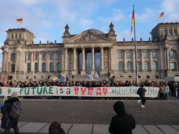 Gruppe von Menschen mit einem Banner mit der Aufschrift "Zukunft ist ein Mensch Neustar ima" vor dem Reichstaggebäude in Berlin, mit seinen architektonischen Details und umgeben von Flaggen unter einem bewölkten Himmel.