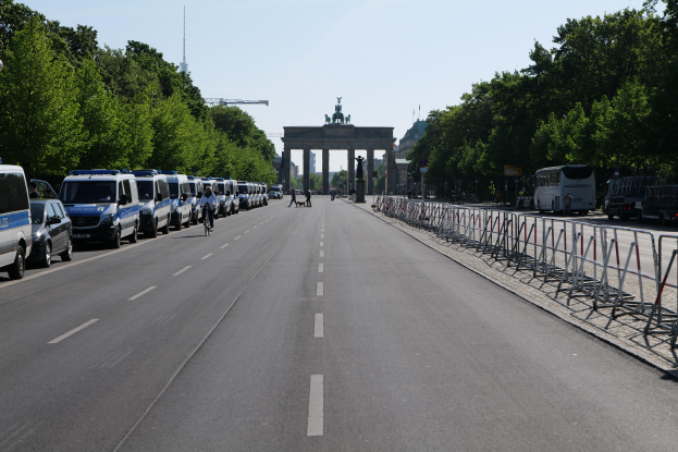 Eine Reihe von Polizeiwagen, die auf der Seite einer Straße vor dem Brandenburger Tor in Berlin, Deutschland, geparkt sind, mit Menschen auf Fahrrädern und auf der Straße, Barrieren und Bäumen an den Seiten und einem Bogen mit Statuen im Hintergrund.