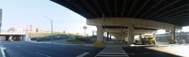 Lastwagen unter einer Brücke auf einer Stadtstraße mit Gebäuden, Straßeninfrastruktur, Fahrzeugen und Grünflächen unter einem klaren blauen Himmel.
