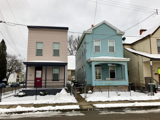 Eine Häuserreihe an einer schneebedeckten Straßenecke mit einem Metallzaun, Bäumen, Fahrzeugen, Pfählen, Drähten und einem bewölkten Himmel.