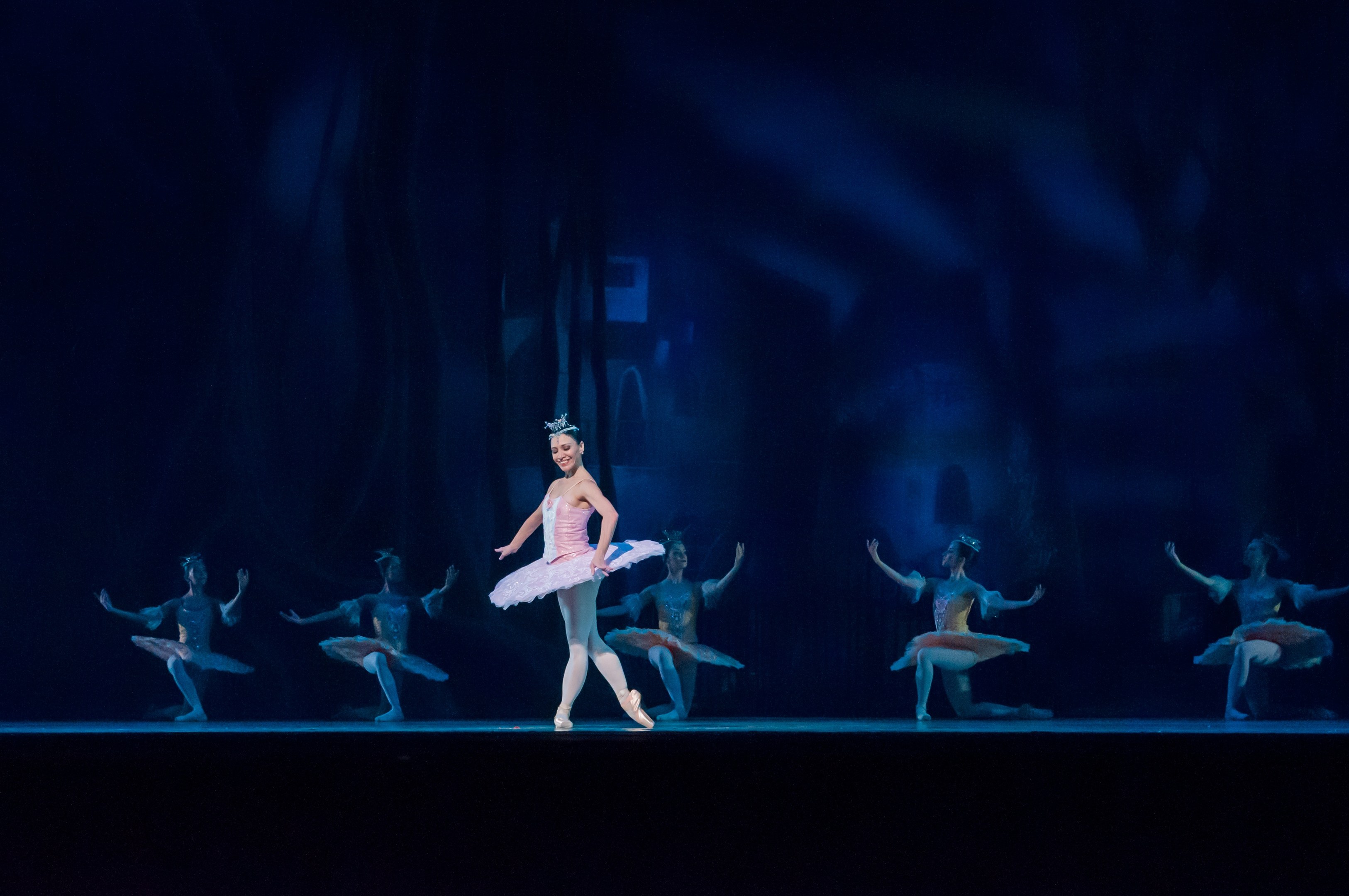 Eine Ballerina in einem pinken Tutu und Spitzenschuhen tanzt auf einer Bühne mit anderen Tänzern in passenden Kostümen vor einem dunklen Hintergrund.