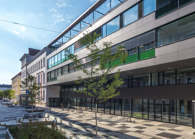 Großes modernes Gebäude mit zahlreichen Fenstern, umgeben von Bäumen und Grünflächen, mit parkenden Fahrzeugen und Fußgängern davor, unter einem bewölkten Himmel.