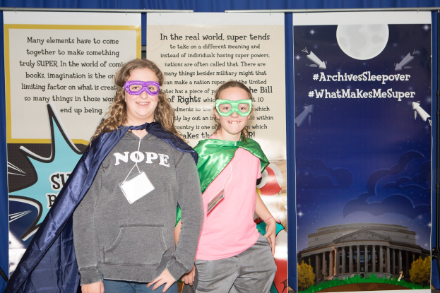 Zwei junge Mädchen in Superheldenkostümen stehen nebeneinander, lächeln hinter Masken, mit einem Vorhanghintergrund und einem Banner mit Bildern eines Gebäudes, Bäume und Himmel.