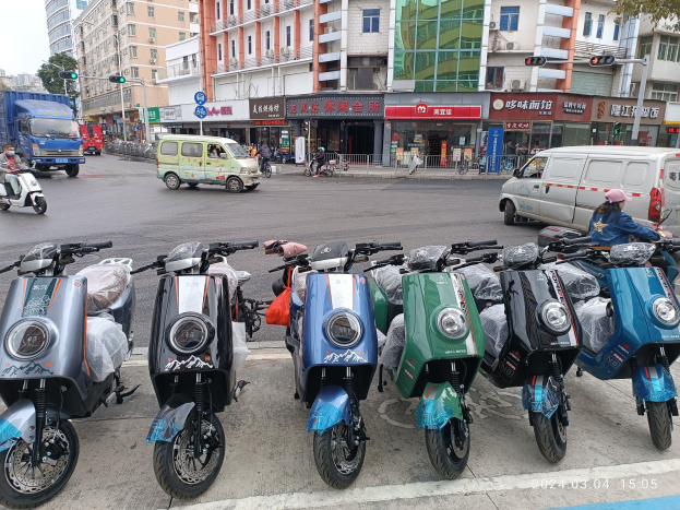 Eine Reihe von Elektroscootern, die auf einer Straße geparkt sind, mit einer Person in einer Mütze davor, Gebäuden, Texttafeln, Verkehrszeichen, Bäumen und einem klaren Himmel im Hintergrund.