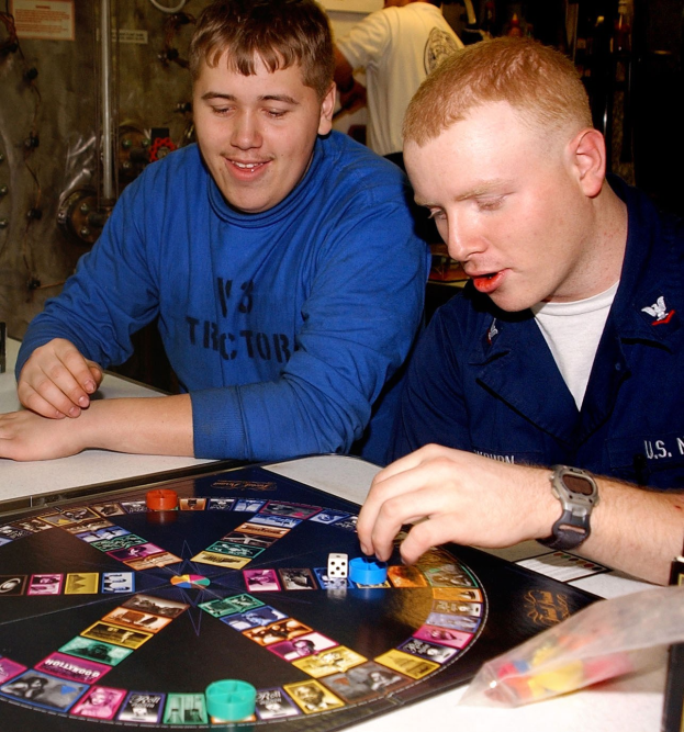 Zwei Männer spielen ein Brettspiel an einem Tisch mit Würfeln und anderen Gegenständen, mit ein paar Leuten im Hintergrund.