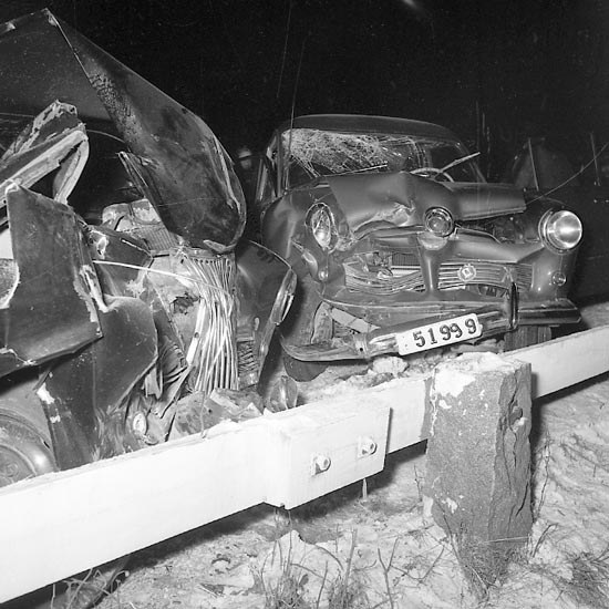 Schwarzes und weißes Foto eines schwer beschädigten Autos mit eingedellter Motorhaube und zersplitterten Fenstern, das am Straßenrand mit Gras und einem Zaun im Hintergrund geparkt ist.