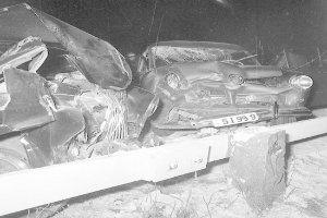 Schwarzes und weißes Foto eines schwer beschädigten Autos mit eingedellter Motorhaube und zersplitterten Fenstern, das am Straßenrand mit Gras und einem Zaun im Hintergrund geparkt ist.