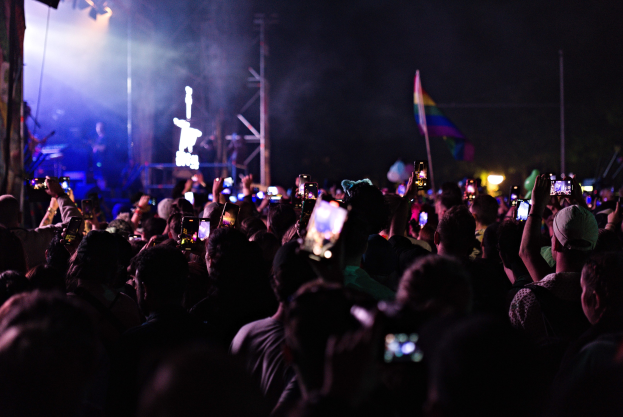 Menge bei einem Konzert mit hochgehaltenen Handys, beleuchtet von Lichtern, umgeben von Pfählen und einer Fahne, während Musiker auf der Bühne spielen.
