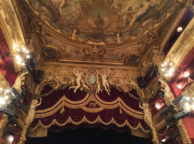 Das Innere eines Theaters mit einem roten Vorhang, Kronleuchtern, Skulpturen, Lichtern und einer Deckenmalerei, das eine Bühne mit ein paar Menschen am Boden zeigt.