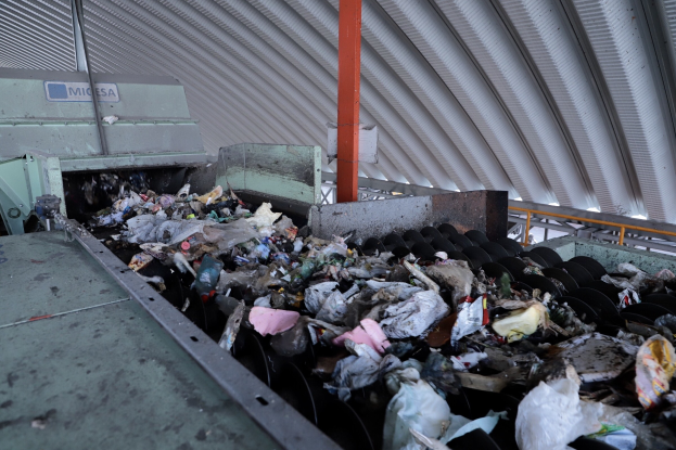 Großer Müllhaufen auf einem Förderband in einer Recyclinganlage mit einer Maschine im Vordergrund und einem Metalldach darüber.