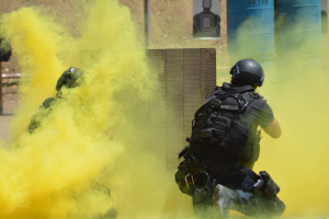 Zwei Polizisten in Einsatzausrüstung mit gelbem Rauch aus dem Mund, vor einer Wand mit einer Tafel und anderen Gegenständen stehend und den Boden unten sichtbar.