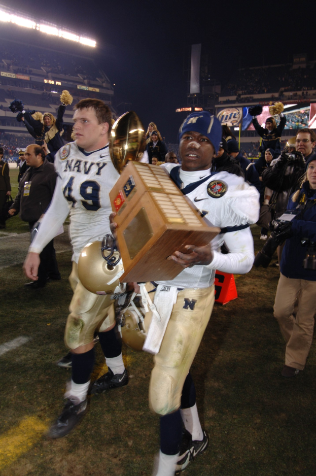 Ein Fussballspieler mit einem Pokal auf einem Feld, umgeben von einer Menge in Sportkleidung, mit Schildern, Lichtern und einem klaren blauen Himmel im Hintergrund.