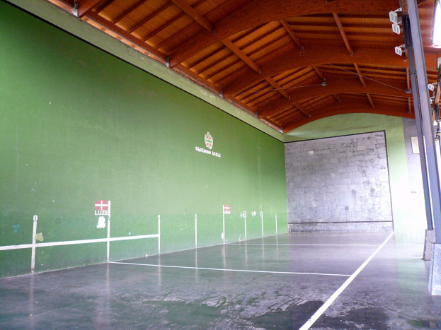 Gro├če Indoor-Tennishalle mit grünen W├Ąnden, Holzdecke, einem Pfosten und deckenmontierten Lichtern auf der rechten Seite.