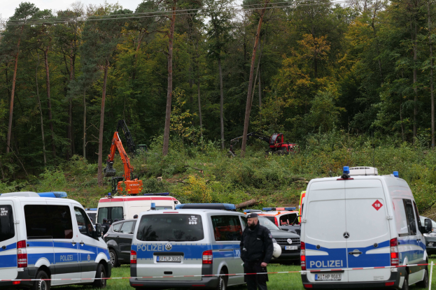 Eine Gruppe von Polizeiwagen auf einem Parkplatz geparkt mit einer Person in der Nähe, Bäume und ein Bagger im Hintergrund unter einem sichtbaren Himmel.