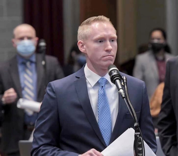 Ein Mann in Anzug und Krawatte spricht an einem Podium mit Papieren vor einem Mikrofon, während andere in der Nähe stehen und eine Vorhangwand im Hintergrund zu sehen ist.