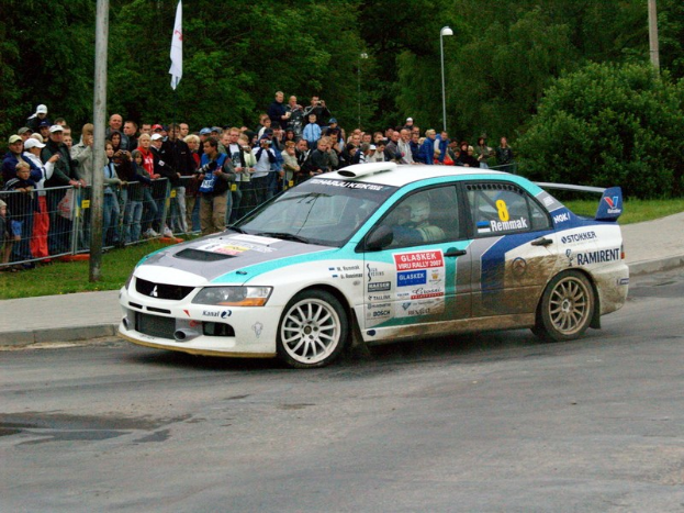 Ein Rallyeauto mit Text und Logos fährt an einer Menge vorbei, die Kameras hält, mit Geländern, Polen mit Flaggen und Bäumen im Hintergrund.