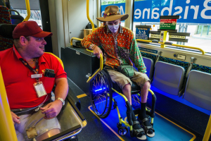 Zwei Männer im Rollstuhl in einem Bus, einer mit roter Mötte und Kappe, der andere mit orangefarbenem Hemd und brauner Mötte, beide mit Masken und Ausweisen, mit gelben Stäben und Glasfenstern, die Bäume, Gebäude und den Himmel zeigen.