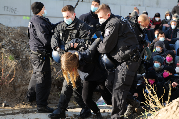 Gruppe von maskierten und gekappten Personen, einige mit Waffen, umringt eine Frau, die von der Polizei eskortiert wird, vor einer Wand mit Text, Pflanzen und Steinen in einer städtischen Umgebung.