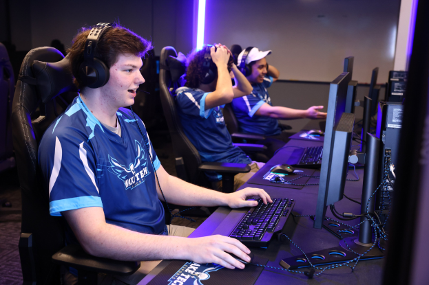 Gruppe junger Männer mit Headsets, die an Computern mit Monitoren, Tastaturen und Mäusen auf einem Schreibtisch spielen, mit beleuchteter Wand im Hintergrund.