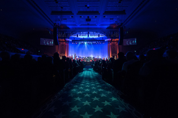 Ein großer Saal mit sitzenden Zuschauern, eine Bühne mit Musikern und Deckenbeleuchtung und -fernseher, was auf ein Konzerthaus während der Nationalkonvention 2019 hinweist.