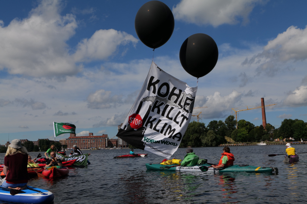 Gruppe von Menschen in Kajaks auf dem Wasser mit einem Banner, das "Kohle Kill Klima" und Paddel hält, umgeben von Bäumen, Gebäuden, Kränen und einem klaren Himmel.