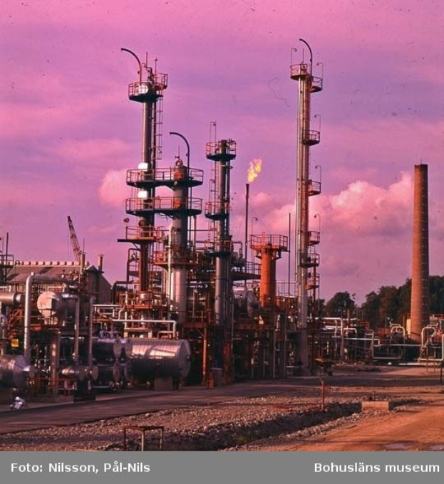 Komplexe industrielle Ölraffinerie mit ausgedehnten Rohrleitungen, Lagertanks, Stützstrukturen, einem entfernten Gebäude und einem Kran unter einem bewölkten Himmel, mit Text unten.