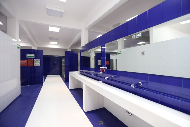 Öffentliches WC mit blauen Kacheln, weißen Tresen, Waschbecken mit Wasserhähnen, einer Plattform mit einer Flasche und anderen Gegenständen, Texttafeln an den Wänden, Deckenleuchten und Stützpfeilern.